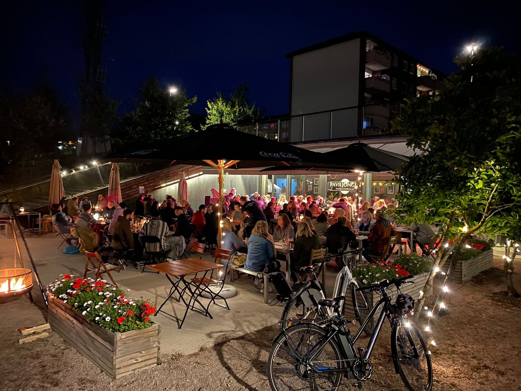 Uteserveringen til Paviljongen i Bjerkedalen park en sommerkveld: fulle bord under parasollene, lysslynger i trærne og bålpanne som brenner.